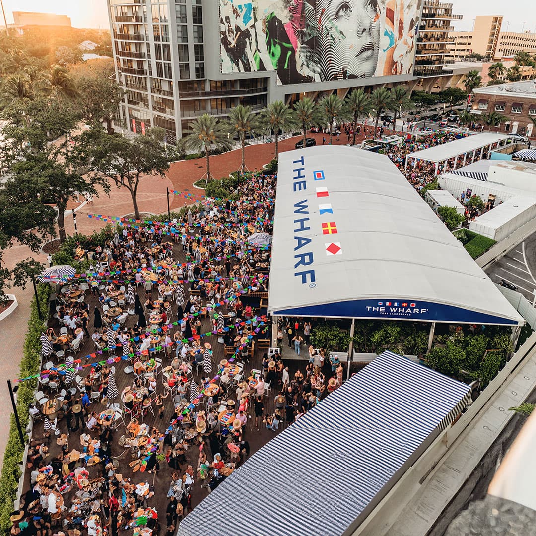 Aerial view of The Wharf Fort Lauderdale event with crowds under colorful bunting and the signature white tent